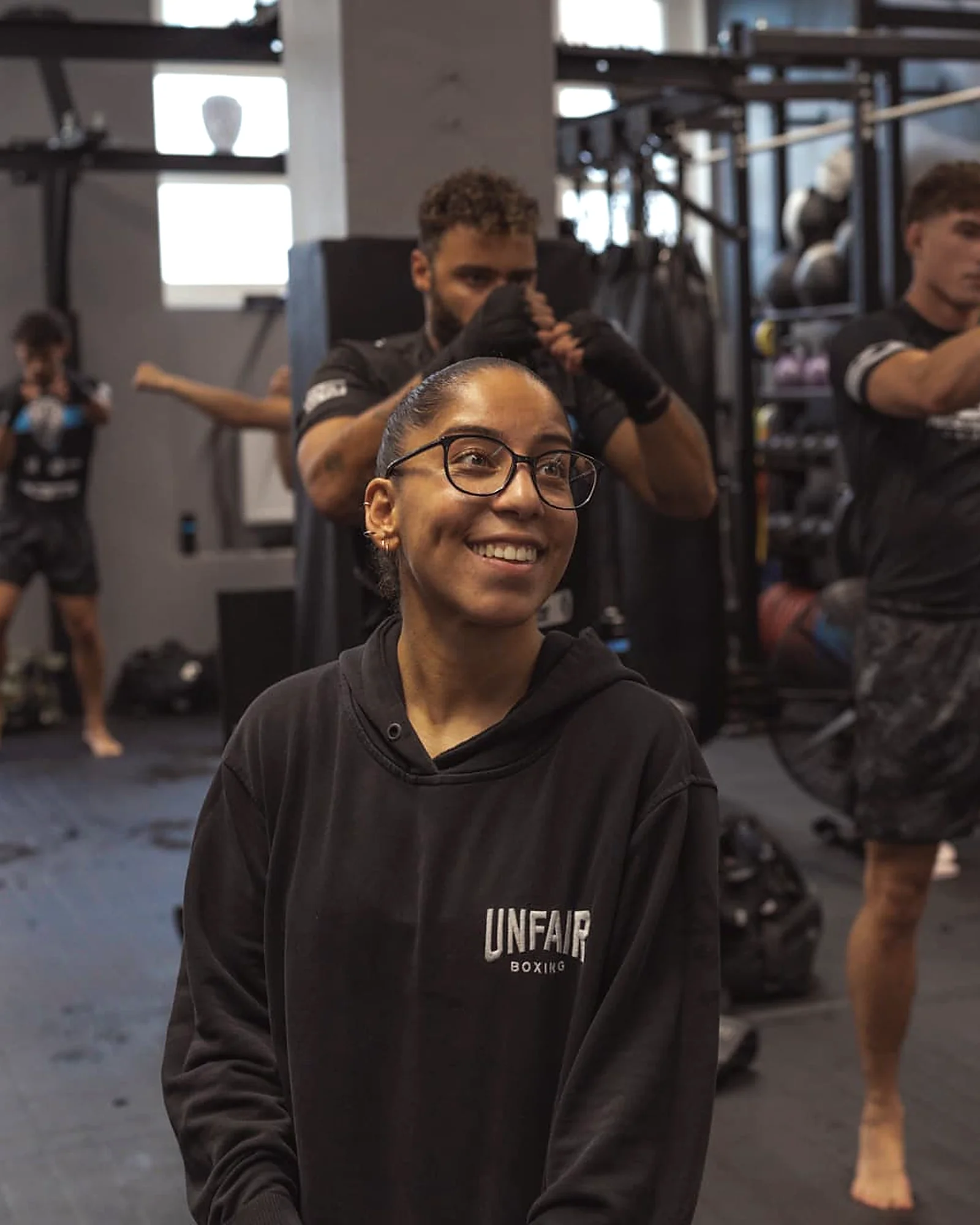 Trainerin Narin lacht in die Kamera, im Hintergrund trainieren Teilnehmerinnen des Ladies-Kurses an Pratzen in der Halle der Barmbek Basch Fightschool